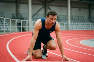 Athlète masculin en position de départ sur une piste d'athlétisme en salle, regard concentré vers l'avant illustrant la vision comme facteur de performance